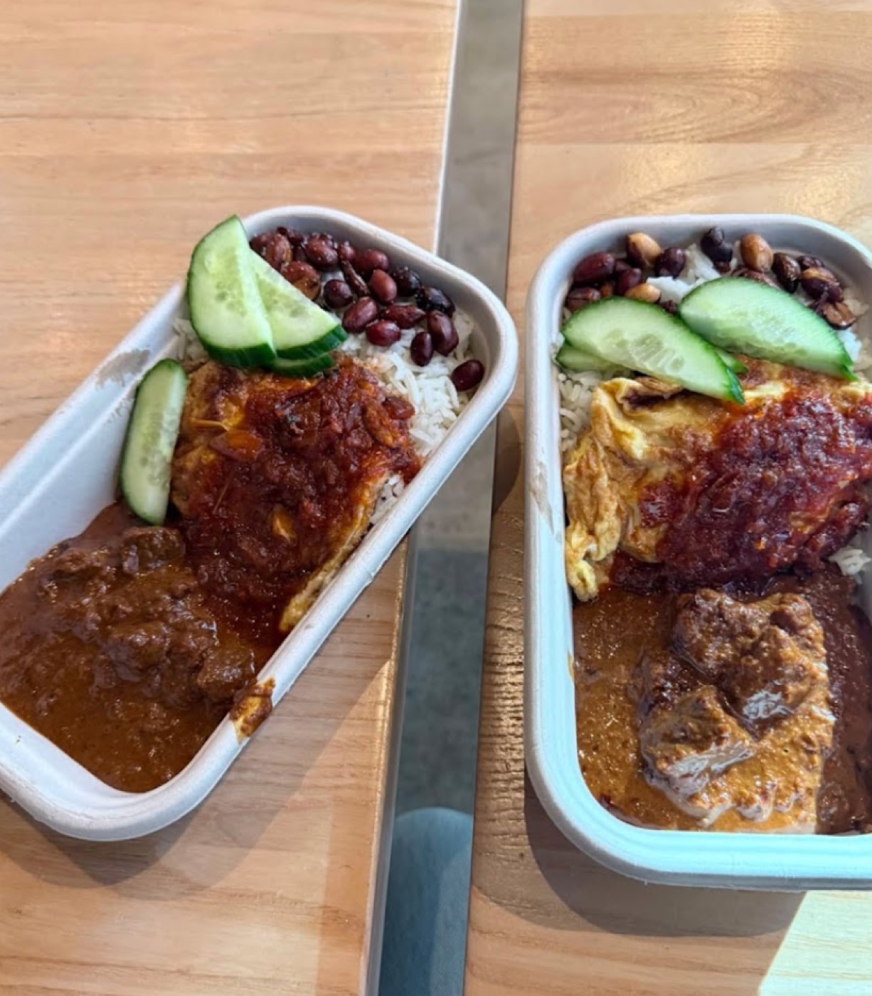 Two nasi lemak takeaway boxes with sambal, beef rendang, cucumber and peanuts